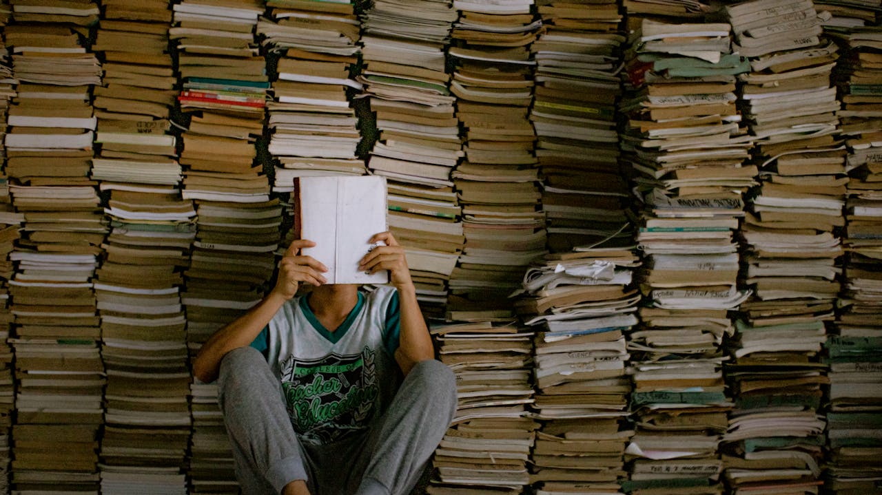 Home A person sitting with a book in front of stacks of books, depicting a library or bookstore atmosphere.